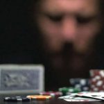 Feszült középkorú férfi öltönyben, sötét szobában pókerasztalnál ül, előtte zsetonok és kártyák, arcán látható a szerencsejáték-függőség miatti stressz és elkeseredettség.