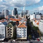 Panorámakép egy vibráló digitális nomád városról 2026-ben: történelmi vörös tetős épületek keverednek modern felhőkarcolókkal, napsütéses égbolt alatt, ideális távmunka és utazás helyszíneként