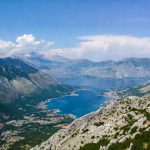 Panorámafotó a Kotori-öbölről a Lovćen-hegyről nézve: mélykék öböl, türkizkék tengerparti részek, meredek sziklás hegyoldalak és zöldellő lejtők, háttérben a montenegrói hegyvonulatok, kék ég enyhén gomolyfelhőkkel.