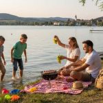 Vidám család kempingezik és piknikezik egy tó partján, Magyarországon. Egy férfi, egy nő és két fiú gyermek látható, a háttérben egy kisváros templomtornyával.